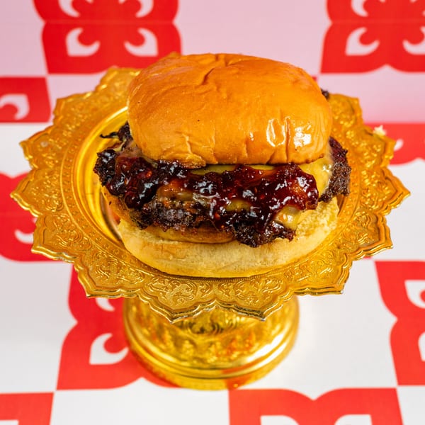 A Ban Ban Burger with cheese and dark, sticky sauce on a golden pedestal against a red and white background.